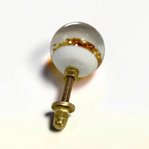 K&ouml;nnte beinhalten: Ein kugelf&ouml;rmiger M&ouml;belknopf mit einer klaren Glasschicht &uuml;ber einer wei&szlig;en Basis, in der bernsteinfarbene Flocken eingebettet sind. Der Knopf ist an einem mit Gewinde versehenen, messingfarbenen Metallbolzen mit Mutter und Unterlegscheibe befestigt.