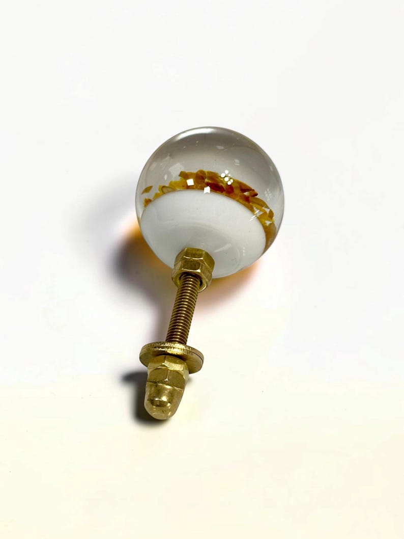 K&ouml;nnte beinhalten: Ein kugelf&ouml;rmiger M&ouml;belknopf mit einer klaren Glasschicht &uuml;ber einer wei&szlig;en Basis, in der bernsteinfarbene Flocken eingebettet sind. Der Knopf ist an einem mit Gewinde versehenen, messingfarbenen Metallbolzen mit Mutter und Unterlegscheibe befestigt.