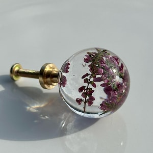 May include: A spherical clear resin cabinet knob featuring embedded purple heather flowers. The knob is attached to a gold-toned metal base and a threaded screw, providing a functional hardware piece for furniture or cabinetry doors and drawers.
