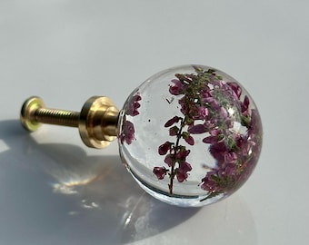 Pode incluir: Um puxador esférico de resina transparente com flores de urze roxas incrustadas. O puxador está fixado a um parafuso e base de metal dourado, formando uma peça de ferragem funcional com um design transparente e arredondado.