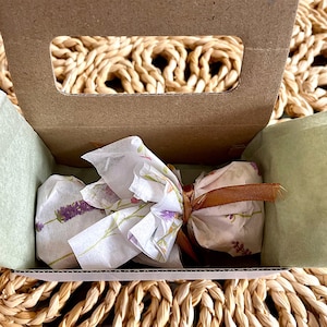 May include: A small brown cardboard box with a handle cutout containing a wrapped item. The item is bundled in white tissue paper featuring a lavender floral print, secured with a thin brown ribbon tied in a bow at the center.