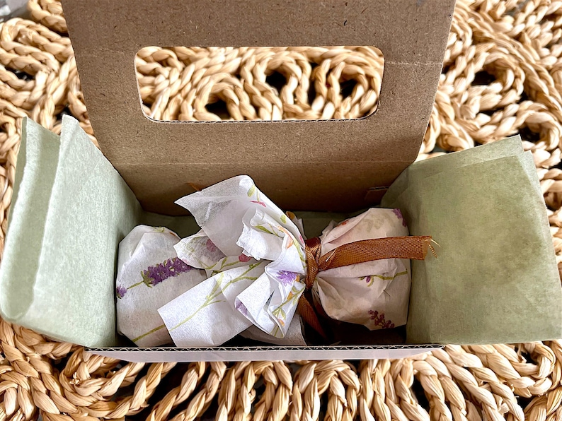 May include: A small brown cardboard box with a handle cutout containing a wrapped item. The item is bundled in white tissue paper featuring a lavender floral print, secured with a thin brown ribbon tied in a bow at the center.