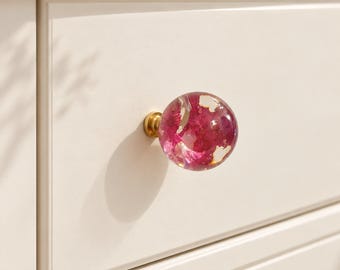 Pode incluir: Um puxador de gaveta esférico com acabamento em resina transparente e elementos florais magenta vibrantes incrustados, fixado a uma base metálica em tom de latão, montado numa frente de gaveta de madeira esbranquiçada.