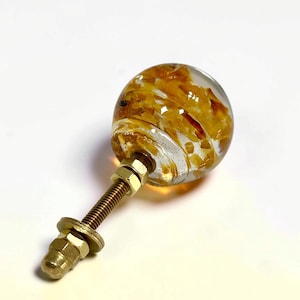 Puede incluir: Un pomo esf&eacute;rico para muebles hecho de resina transparente con incrustaciones de escamas color &aacute;mbar. El pomo est&aacute; unido a un perno de metal roscado en tono lat&oacute;n con dos tuercas hexagonales a juego y una arandela de montaje.
