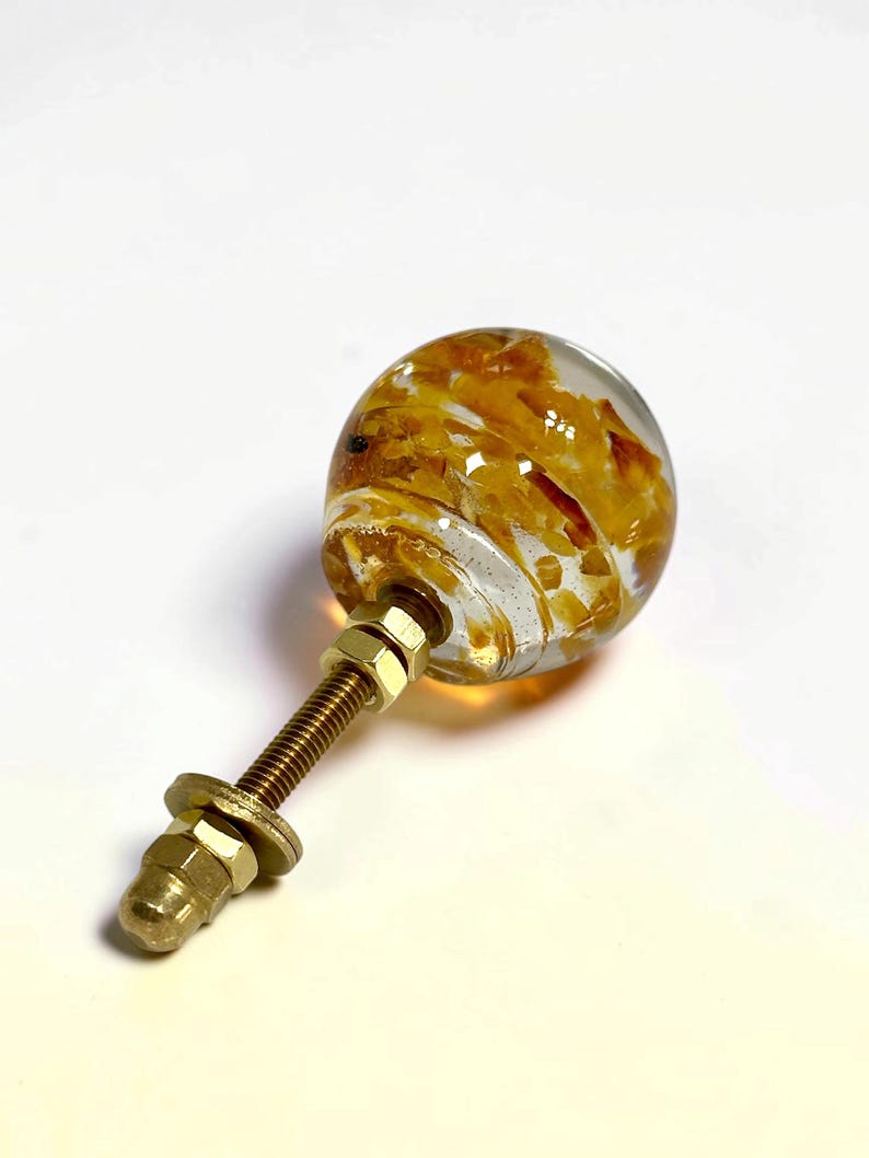 Puede incluir: Un pomo esf&eacute;rico para muebles hecho de resina transparente con incrustaciones de escamas color &aacute;mbar. El pomo est&aacute; unido a un perno de metal roscado en tono lat&oacute;n con dos tuercas hexagonales a juego y una arandela de montaje.