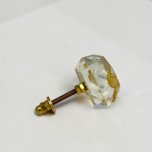 May include: A clear, faceted, geometric cabinet knob made of resin with embedded gold-colored leaf flakes. The knob features a gold-toned metal bolt extending from the back, complete with a matching gold-colored nut and washer for secure installation.
