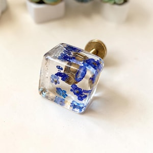 May include: A square-shaped cabinet knob made of clear resin, featuring embedded small blue flowers throughout the transparent material. The knob is attached to a circular brass-coloured metal base and mounting hardware extending from the back.