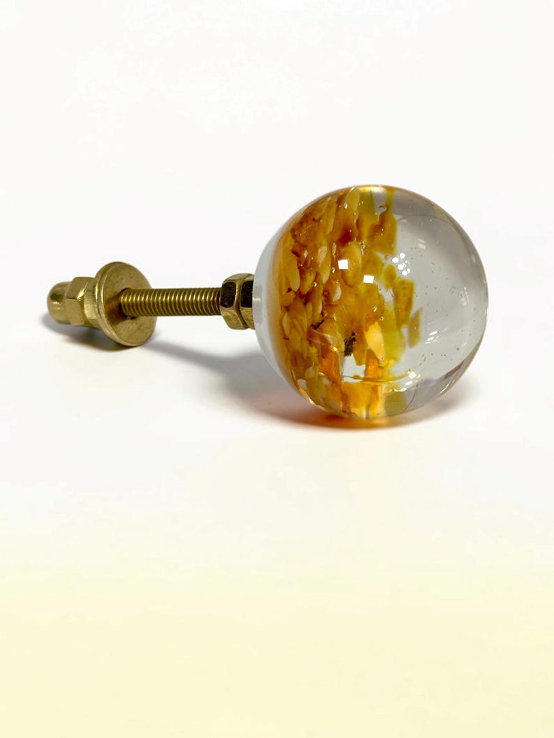 K&ouml;nnte beinhalten: Ein kugelf&ouml;rmiger M&ouml;belknopf aus klarem Glas mit einem eingebetteten gelben und orangefarbenen Texturmuster. Der Knopf verf&uuml;gt &uuml;ber einen metallischen, goldfarbenen Gewindebolzen, eine Unterlegscheibe und eine Mutter zur Montage.