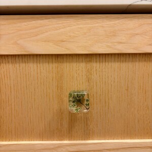 Puede incluir: Un tirador de mueble cuadrado de resina transparente con flores blancas secas y follaje verde incrustados. El tirador est&aacute; montado en un frente de caj&oacute;n de madera natural de tono claro con veta visible, bajo una encimera blanca.