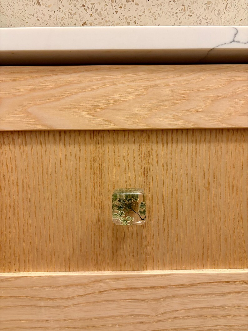 Puede incluir: Un tirador de mueble cuadrado de resina transparente con flores blancas secas y follaje verde incrustados. El tirador est&aacute; montado en un frente de caj&oacute;n de madera natural de tono claro con veta visible, bajo una encimera blanca.