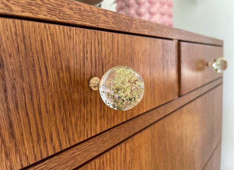 Pode incluir: Um puxador de gaveta redondo em resina transparente com flores secas brancas e verdes embutidas, fixado a uma gaveta de madeira com uma base de metal dourado. O puxador tem uma forma esf&eacute;rica lisa e est&aacute; montado numa superf&iacute;cie de madeira.