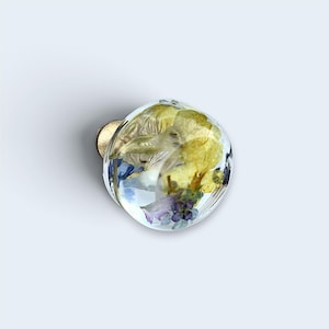 May include: A round cabinet knob made of clear resin, featuring embedded dried yellow and purple flowers inside the dome. The knob has a metallic gold-toned base attachment visible at the back, providing a smooth, spherical surface.