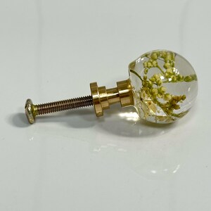 Pode incluir: Um puxador de m&oacute;vel com uma cabe&ccedil;a esf&eacute;rica de resina transparente contendo pequenas flores secas amarelo-esverdeadas. O puxador est&aacute; fixado a uma base de metal dourado e a um parafuso roscado, sendo uma pe&ccedil;a de ferragem funcional.