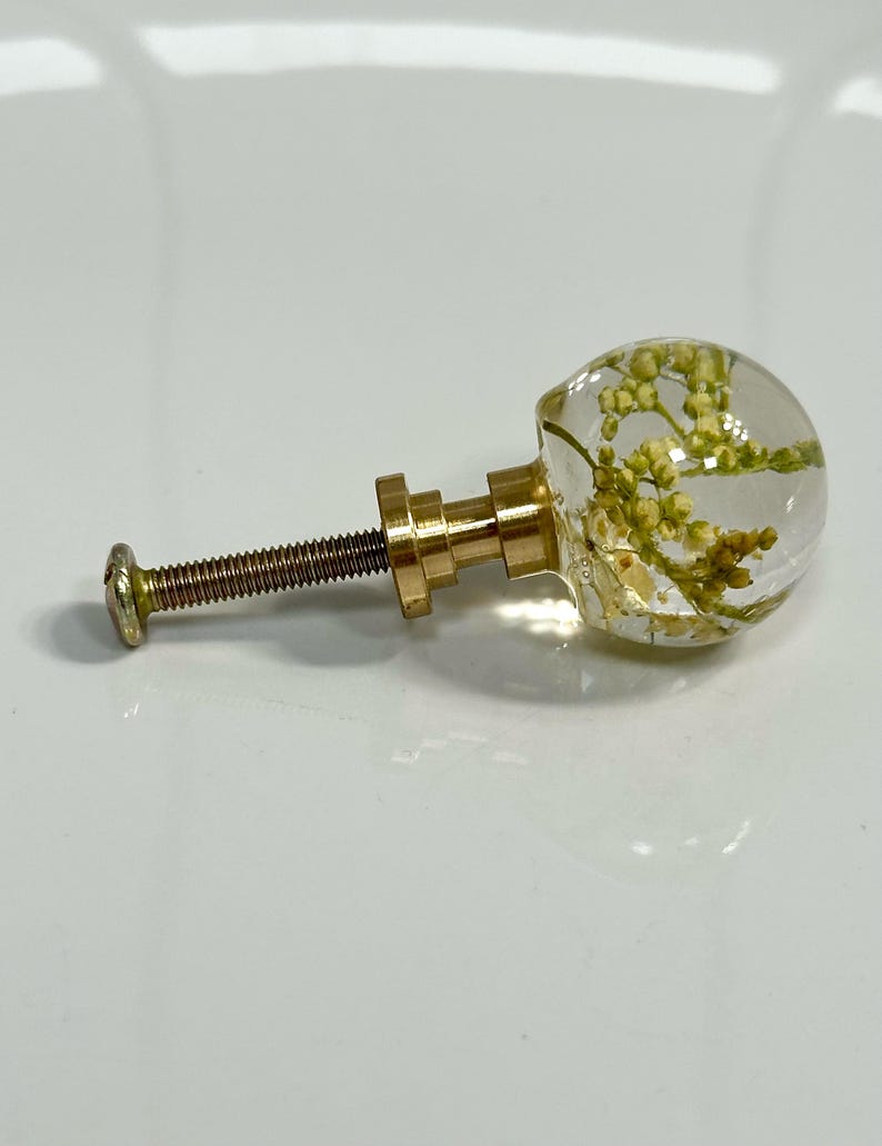 Pode incluir: Um puxador de m&oacute;vel com uma cabe&ccedil;a esf&eacute;rica de resina transparente contendo pequenas flores secas amarelo-esverdeadas. O puxador est&aacute; fixado a uma base de metal dourado e a um parafuso roscado, sendo uma pe&ccedil;a de ferragem funcional.