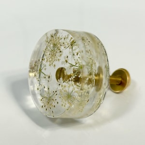 Puede incluir: Un pomo redondo para armario hecho de resina transparente con flores blancas prensadas incrustadas. El pomo cuenta con un tornillo de metal dorado y una fijaci&oacute;n que sobresale por detr&aacute;s, ofreciendo un punto de montaje funcional.