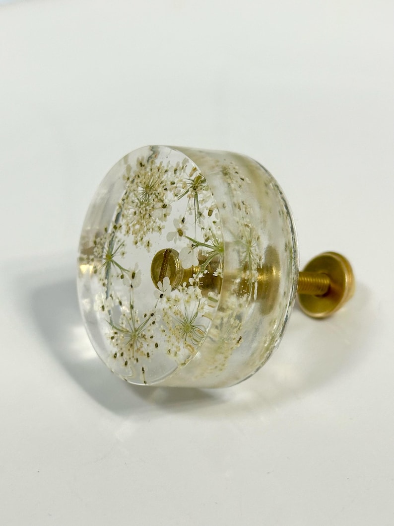 Puede incluir: Un pomo redondo para armario hecho de resina transparente con flores blancas prensadas incrustadas. El pomo cuenta con un tornillo de metal dorado y una fijaci&oacute;n que sobresale por detr&aacute;s, ofreciendo un punto de montaje funcional.