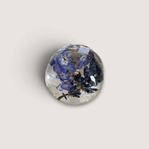 May include: A spherical clear resin bead containing preserved blue forget-me-not flowers and small dark blue buds. The transparent material creates a smooth, glossy surface that encases the botanical elements in a rounded, three-dimensional form.