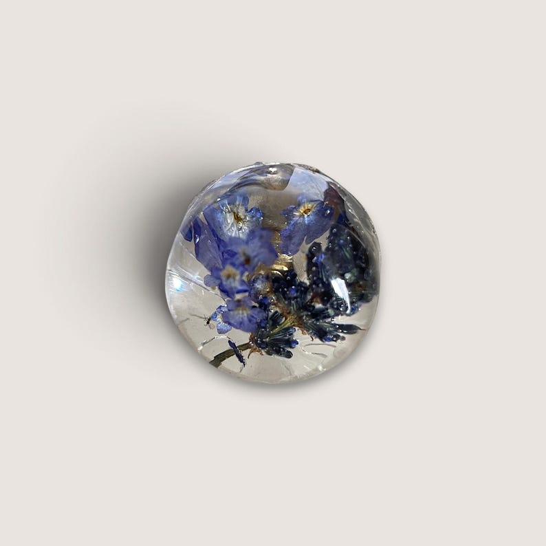 May include: A spherical clear resin bead containing preserved blue forget-me-not flowers and small dark blue buds. The transparent material creates a smooth, glossy surface that encases the botanical elements in a rounded, three-dimensional form.