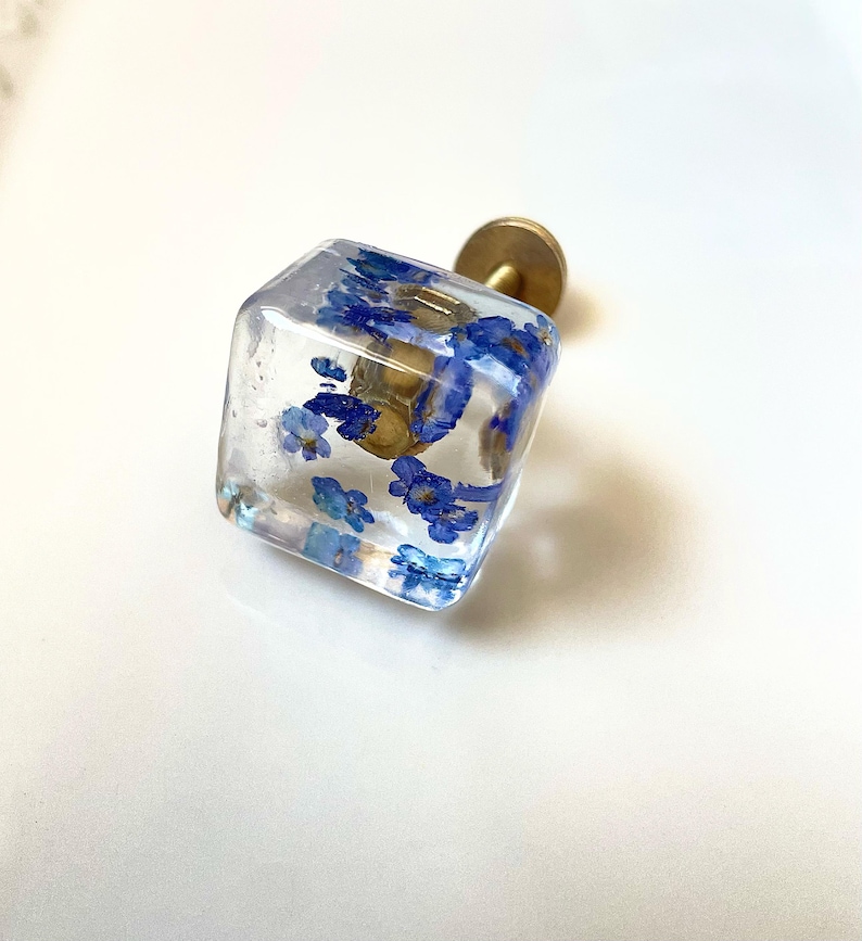 May include: A square-shaped cabinet knob made of clear resin with small, dried blue flowers embedded inside. The knob features a metallic gold-toned circular base and a short, cylindrical mounting post extending from the back of the square piece.