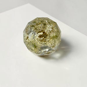May include: A clear, multifaceted resin sphere containing embedded white dried flowers and small gold-colored glitter particles. The object has a geometric, diamond-like cut surface that reflects light, showcasing the botanical elements inside.