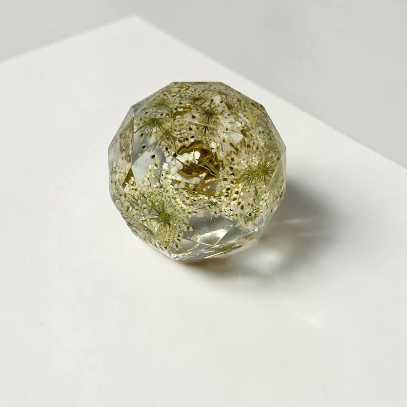 May include: A clear, multifaceted resin sphere containing embedded white dried flowers and small gold-colored glitter particles. The object has a geometric, diamond-like cut surface that reflects light, showcasing the botanical elements inside.