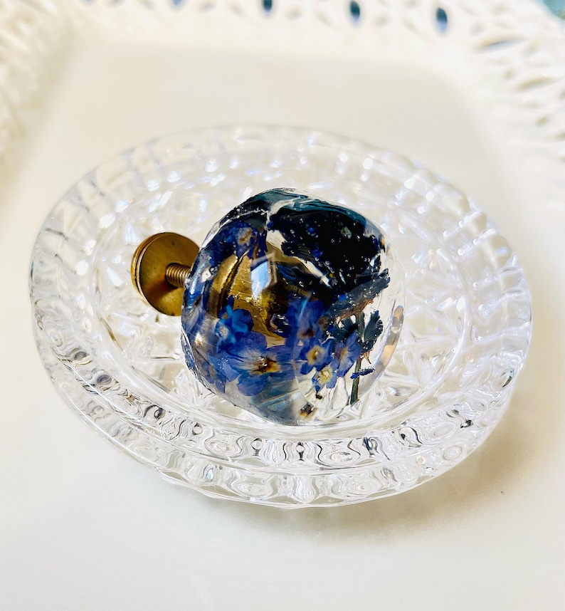 May include: A round cabinet knob made of clear resin containing embedded blue forget-me-not flowers and gold leaf flakes. The knob features a brass-colored metal screw base extending from the back, designed for attachment to furniture or drawers.