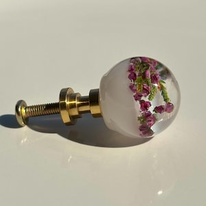 Può includere: Un pomello sferico per mobili con una cupola in resina trasparente che racchiude piccoli fiori di erica viola e fogliame verde. Il pomello è fissato a una base in metallo color oro e a una vite filettata, creando un elemento di ferramenta.