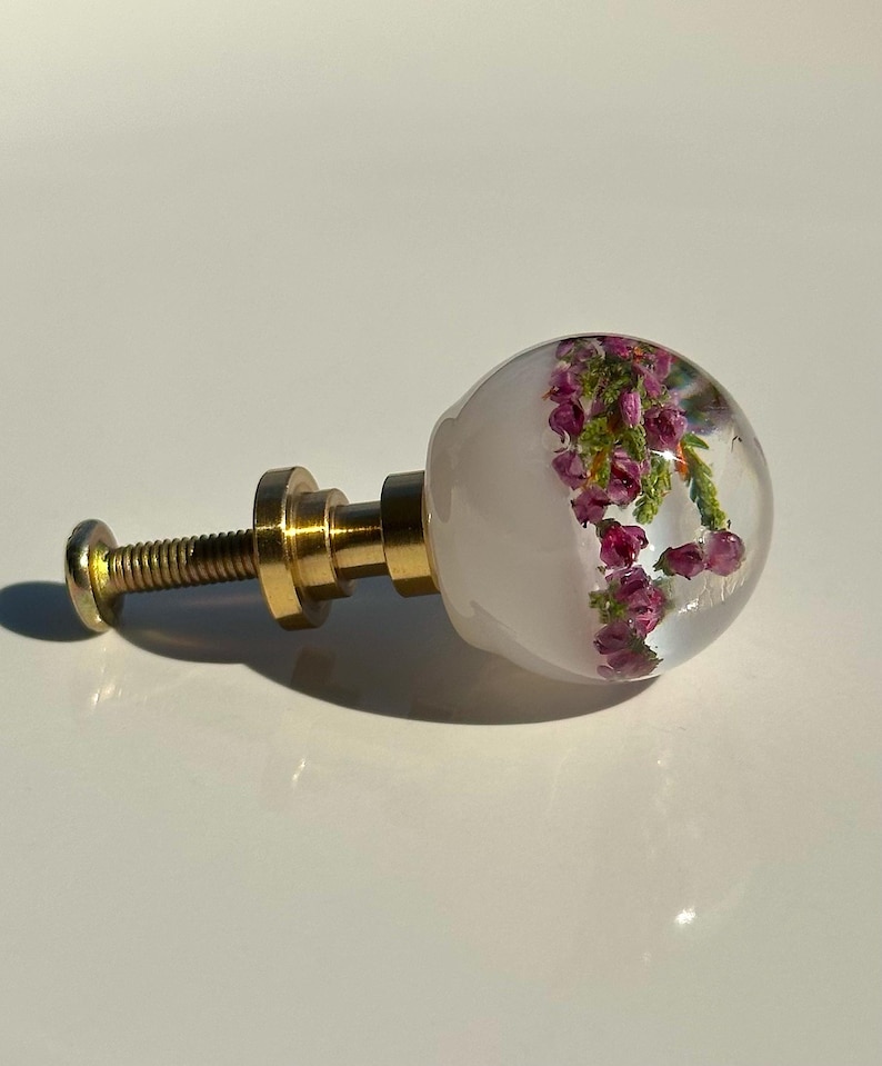 May include: A spherical cabinet knob featuring a clear resin dome embedded with small purple heather flowers and green foliage. The knob is attached to a metallic gold-toned base and a threaded screw, providing a functional hardware component.