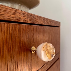 Può includere: Un pomello per mobili rotondo con una base bianca opaca e liscia e una parte superiore trasparente a cupola contenente scaglie di foglia d'oro incastonate. Il pomello è fissato al frontale di un cassetto da una base cilindrica in ottone dorato.