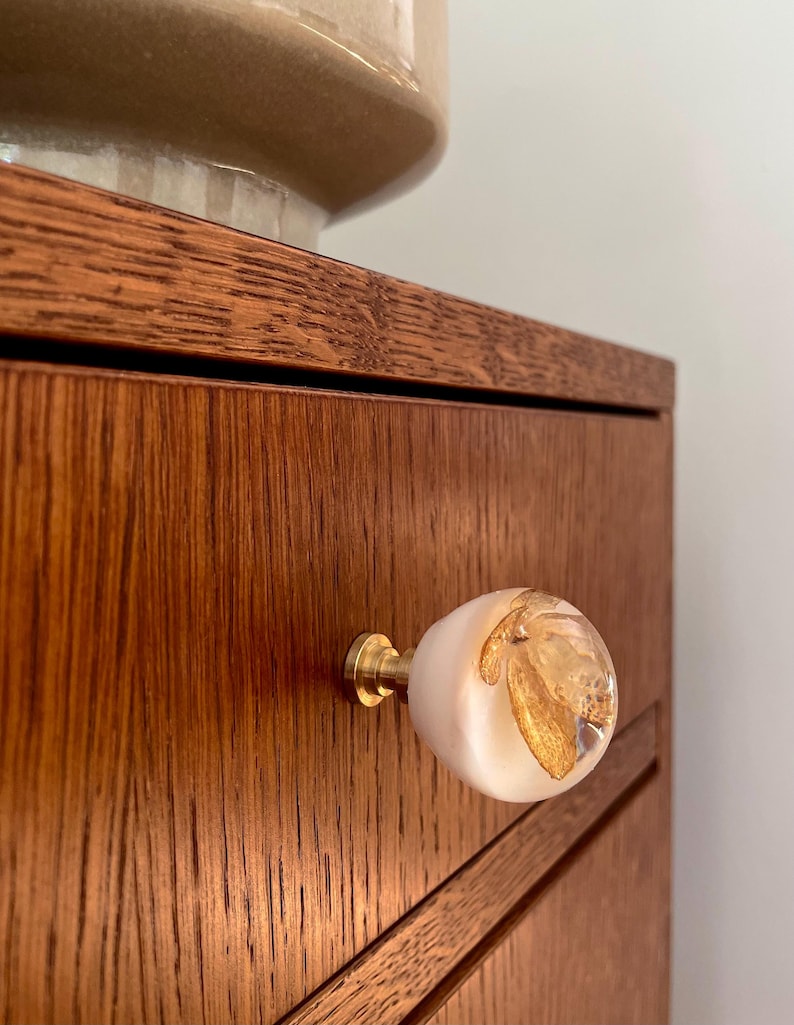 May include: A round cabinet knob featuring a smooth, opaque white base and a clear, dome-shaped top containing embedded gold leaf flakes. The knob is attached to a drawer front by a cylindrical, metallic gold-toned brass hardware base.