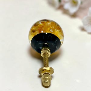 May include: A spherical cabinet knob featuring a clear resin top filled with amber and gold-toned stone fragments, set against a solid black base. The hardware includes a threaded brass-toned metal bolt with two matching nuts and a washer.