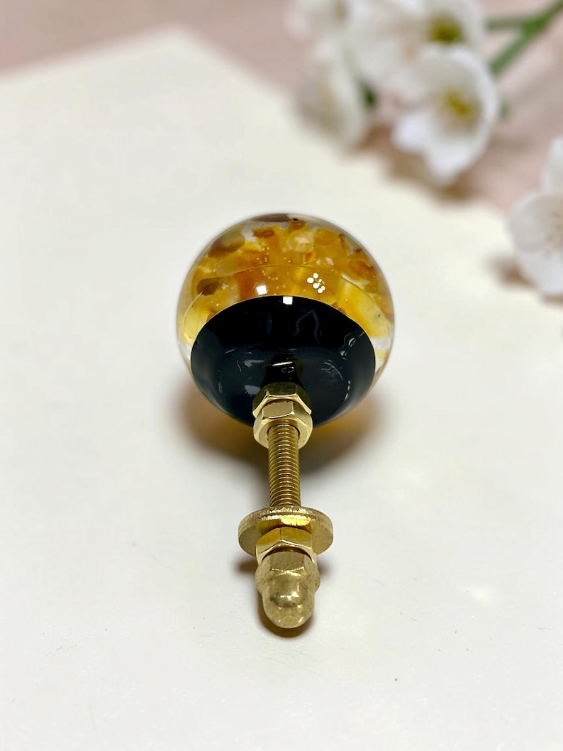 May include: A spherical cabinet knob featuring a clear resin top filled with amber and gold-toned stone fragments, set against a solid black base. The hardware includes a threaded brass-toned metal bolt with two matching nuts and a washer.