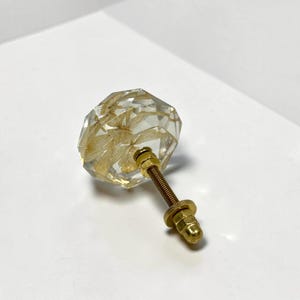 May include: A clear, faceted, octagonal cabinet knob made of resin with embedded dried white flowers inside. The knob features a gold-toned metal threaded bolt, a matching washer, and a hexagonal nut attached to the back for installation.