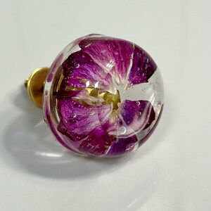 May include: A round cabinet knob featuring a clear resin dome that encapsulates a vibrant purple flower. The floral petals are visible through the transparent material, which is attached to a circular gold-toned metal base for mounting purposes.