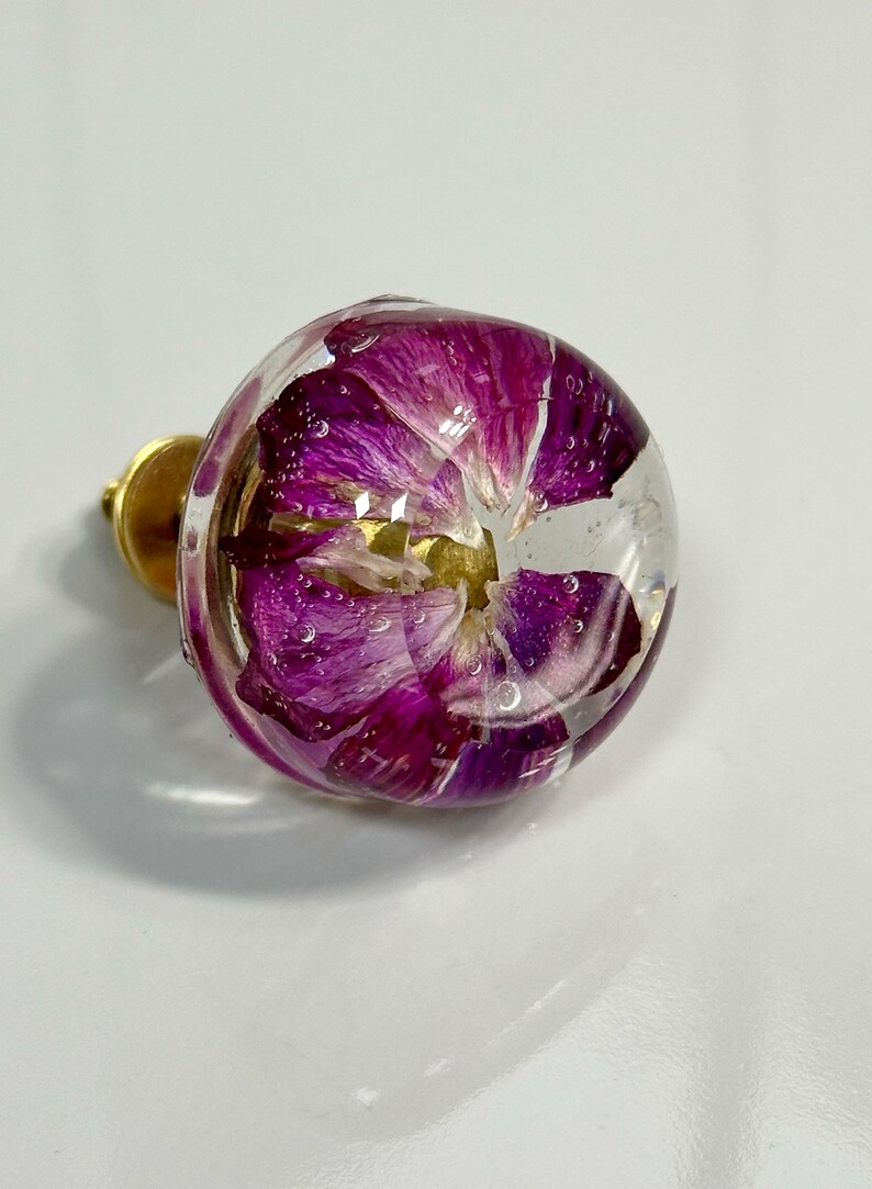 May include: A round cabinet knob featuring a clear resin dome that encapsulates a vibrant purple flower. The floral petals are visible through the transparent material, which is attached to a circular gold-toned metal base for mounting purposes.