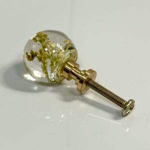Pode incluir: Um puxador esf&eacute;rico para m&oacute;veis com uma c&uacute;pula de resina transparente contendo flores amarelas secas incrustadas. O puxador est&aacute; fixado a uma base de metal dourado com um parafuso roscado, servindo como ferragem para m&oacute;veis ou arm&aacute;rios.