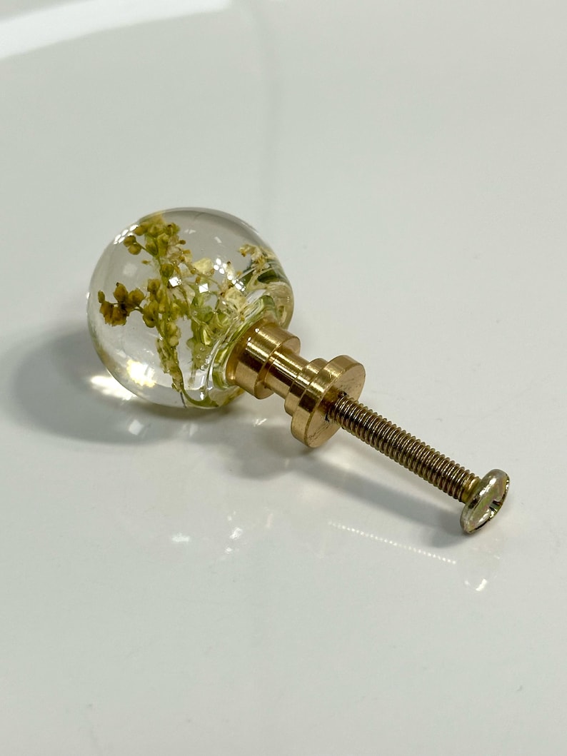 Pode incluir: Um puxador esf&eacute;rico para m&oacute;veis com uma c&uacute;pula de resina transparente contendo flores amarelas secas incrustadas. O puxador est&aacute; fixado a uma base de metal dourado com um parafuso roscado, servindo como ferragem para m&oacute;veis ou arm&aacute;rios.