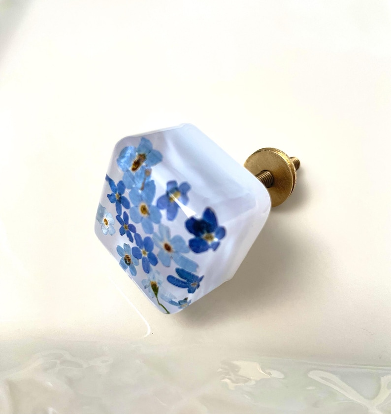 Puede incluir: Un pomo para mueble de forma cuadrada hecho de resina transparente, con peque&ntilde;as flores azules de nomeolvides con centros amarillos incrustadas. El pomo est&aacute; unido a una base met&aacute;lica de color lat&oacute;n con un tornillo roscado en la parte posterior.