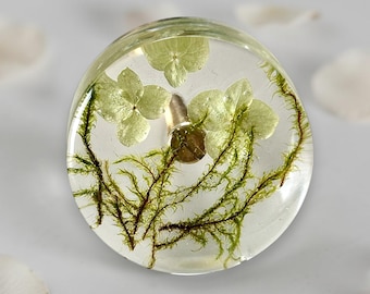 May include: A circular cabinet knob made of clear resin, featuring embedded light green hydrangea petals and dark green moss. A metallic gold-toned screw hardware component is visible in the center of the transparent resin disc.