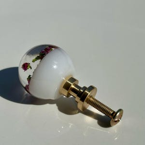 May include: A spherical cabinet knob featuring a clear resin top half embedded with small purple flowers and green foliage, a solid white opaque bottom half, and a gold-toned metal base with a threaded screw for installation and attachment.