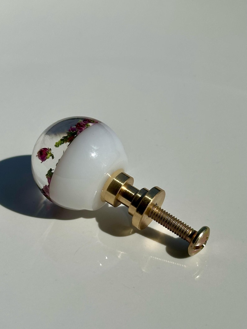 May include: A spherical cabinet knob featuring a clear resin top half embedded with small purple flowers and green foliage, a solid white opaque bottom half, and a gold-toned metal base with a threaded screw for installation and attachment.