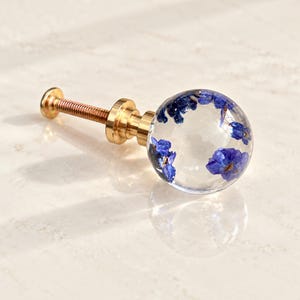 Peut inclure: Un bouton de meuble sphérique composé d'une boule en résine transparente incrustée de petites fleurs bleues séchées. Le bouton est fixé sur une base métallique dorée avec une vis filetée, formant une pièce de quincaillerie fonctionnelle.
