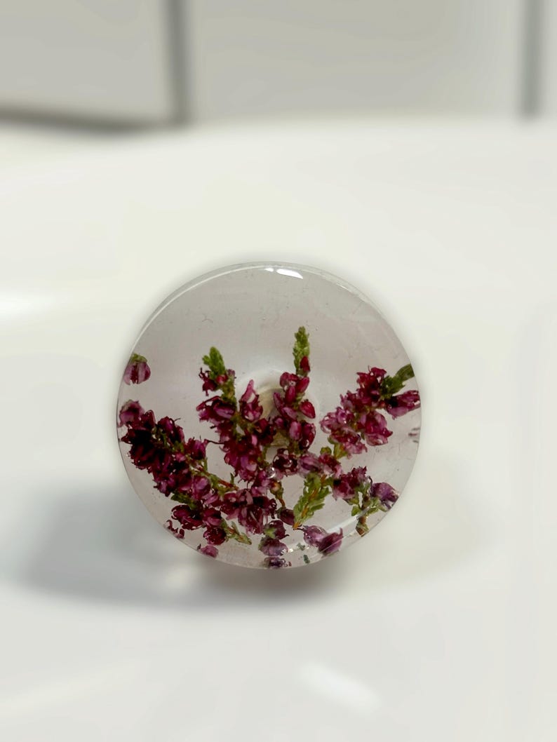 Puede incluir: Un pomo circular con un cuerpo de resina transparente y clara que contiene flores de brezo secas de color p&uacute;rpura y rosa. El pomo tiene una superficie lisa y pulida con una base cil&iacute;ndrica visible en el centro.
