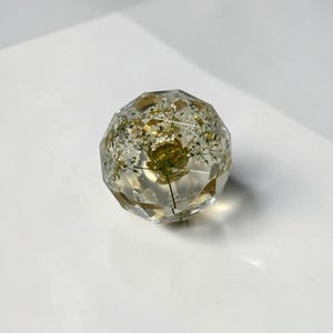 May include: A clear, multi-faceted resin sphere containing delicate, pressed white flowers with green stems. The geometric, crystal-like shape features numerous flat surfaces that catch the light, showcasing the botanical elements embedded within the transparent material.