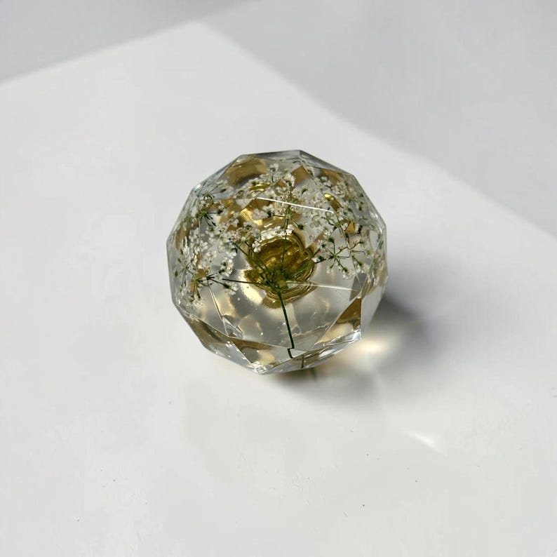 May include: A clear, multi-faceted resin sphere containing delicate, pressed white flowers with green stems. The geometric, crystal-like shape features numerous flat surfaces that catch the light, showcasing the botanical elements embedded within the transparent material.