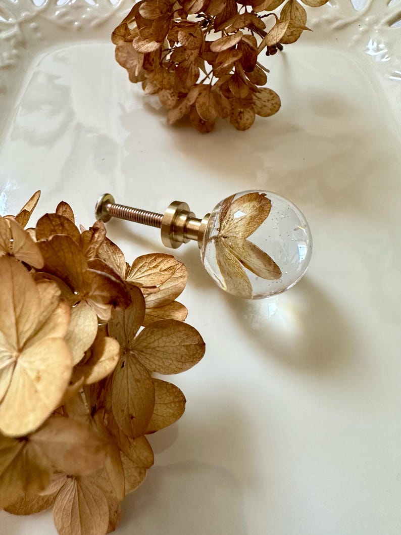Puede incluir: Un pomo esf&eacute;rico para muebles hecho de resina transparente que contiene un p&eacute;talo de flor de hortensia seca de color marr&oacute;n. El pomo cuenta con una base de metal dorado y un tornillo roscado para su fijaci&oacute;n, junto a flores de hortensia secas.