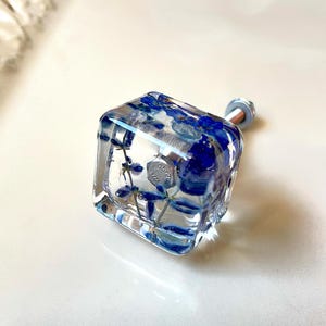 Op de afbeelding: Een vierkante meubelknop van heldere hars met ingebedde gedroogde blauwe bloemen. De knop is voorzien van een metalen zilverkleurige bout en moer als bevestiging, die zichtbaar is door het transparante materiaal van de kubus.