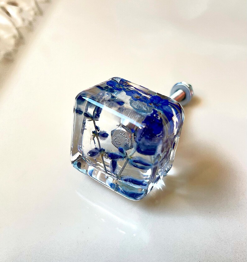 Op de afbeelding: Een vierkante meubelknop van heldere hars met ingebedde gedroogde blauwe bloemen. De knop is voorzien van een metalen zilverkleurige bout en moer als bevestiging, die zichtbaar is door het transparante materiaal van de kubus.