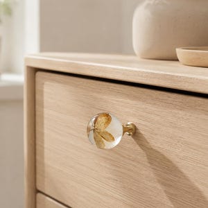 Puede incluir: Un tirador de mueble esf&eacute;rico con un cuerpo de resina transparente que contiene un p&eacute;talo de flor marr&oacute;n seco incrustado, unido a una base met&aacute;lica de tono lat&oacute;n, montado en un frente de caj&oacute;n de madera clara con veta horizontal visible.