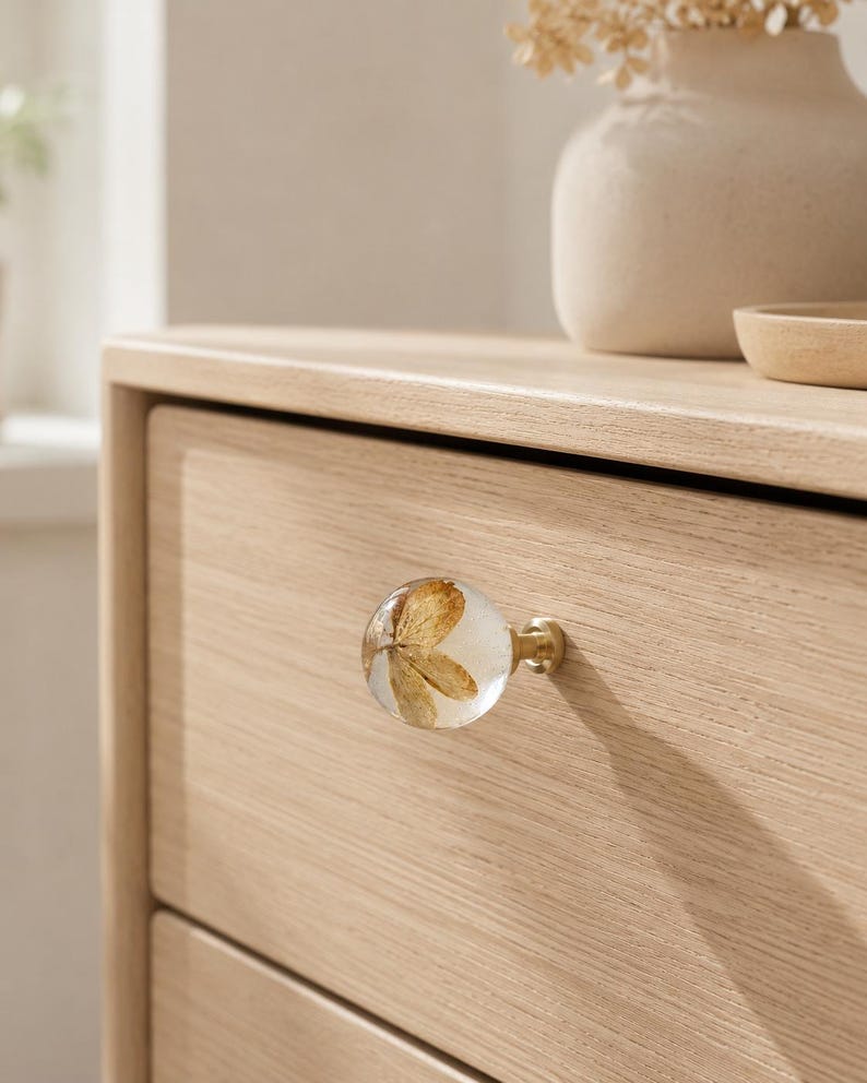 Puede incluir: Un tirador de mueble esf&eacute;rico con un cuerpo de resina transparente que contiene un p&eacute;talo de flor marr&oacute;n seco incrustado, unido a una base met&aacute;lica de tono lat&oacute;n, montado en un frente de caj&oacute;n de madera clara con veta horizontal visible.